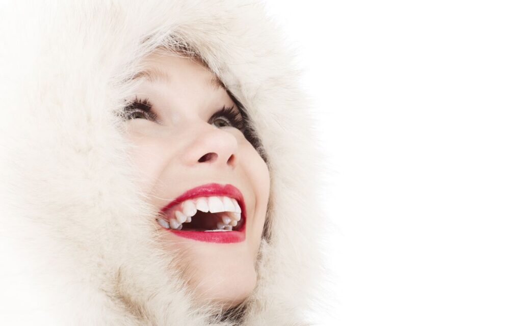 A woman with vibrant red lips and a fur hood smiles joyfully, looking upward.