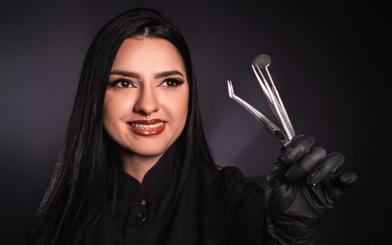 Smiling dentist in black gloves holding dental tools, with a dark background in studio lighting.
