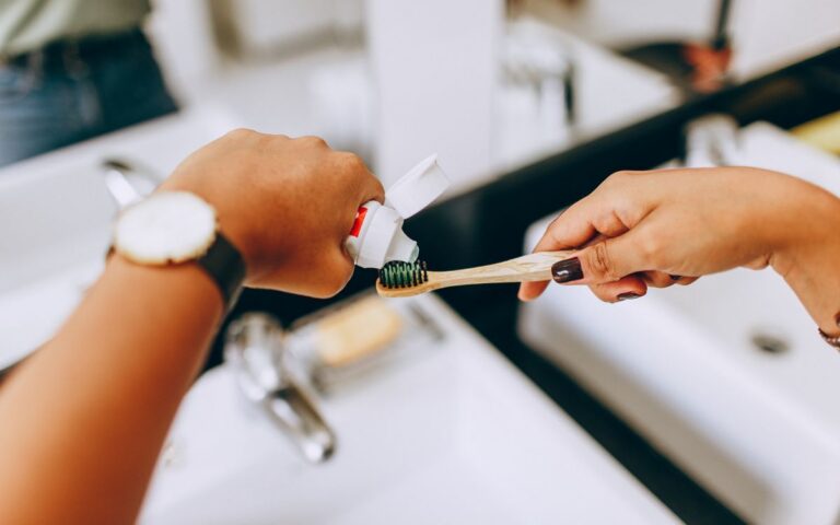 Close-up of a woman putting toothpaste on a bamboo toothbrush in a modern bathroom setting.