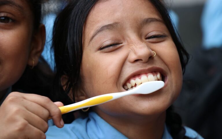 A cheerful child laughing while brushing her teeth with a yellow and white toothbrush.
