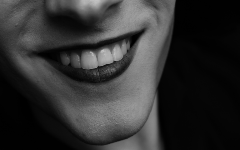 Black and white of unrecognizable person with dark lips and white even teeth smiling on black background