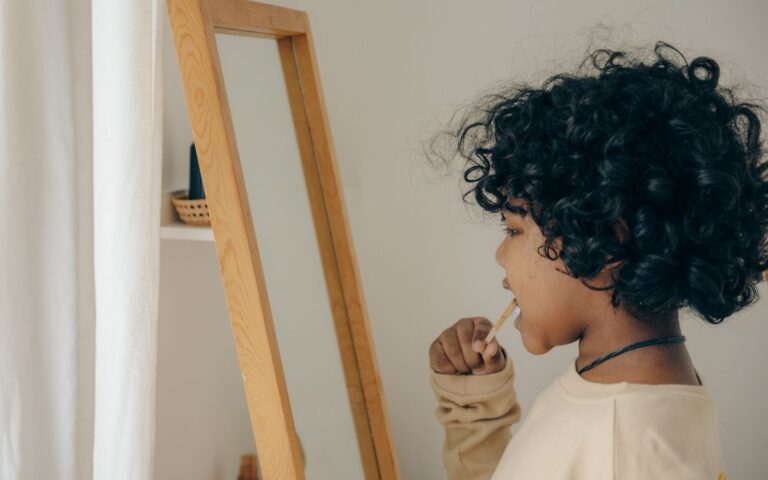 Side view of focused African American child with curly hair standing opposite mirror and brushing teeth while looking at