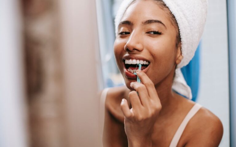 Smiling African American female with white towel on head cleaning teeth with dental flosser in bathroom