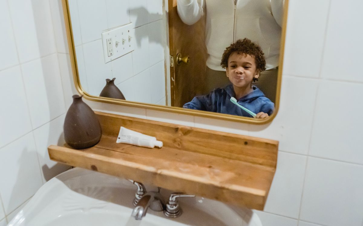 Cheerful African American boy with toothbrush and looking at reflection in mirror while standing in bathroom near sink w