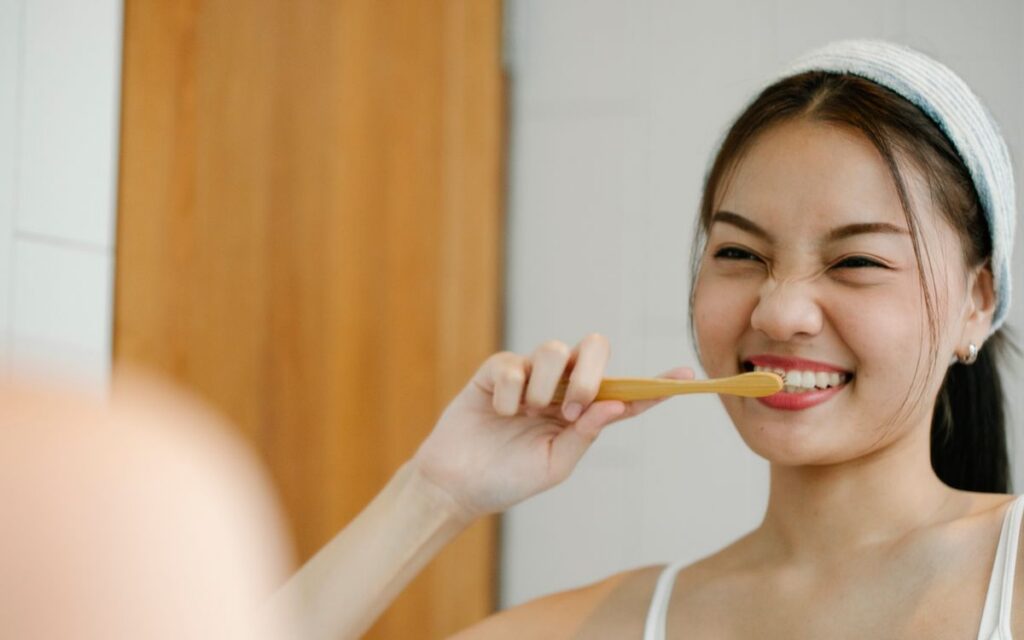 Happy Asian female with black hair brushing teeth with wooden toothbrush while looking in mirror during morning hygienic