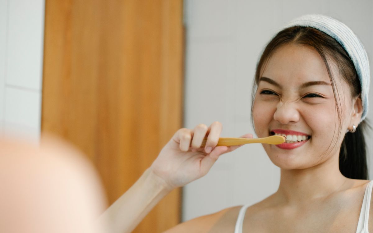 Happy Asian female with black hair brushing teeth with wooden toothbrush while looking in mirror during morning hygienic