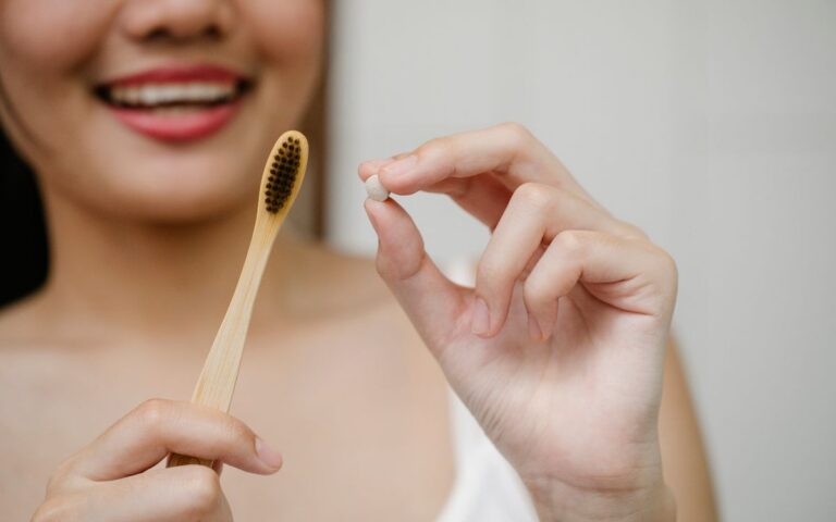 Unrecognizable smiling female with wooden toothbrush and small toothpaste tablet in hand standing in light bathroom duri