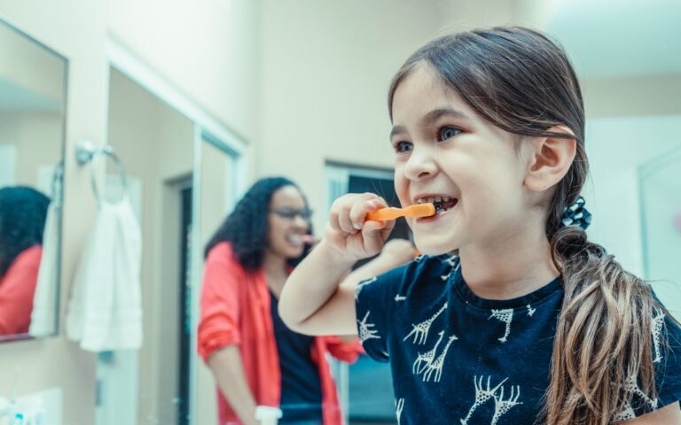 Young girl brushing her teeth in the bathroom with parental supervision, promoting healthy dental habits.