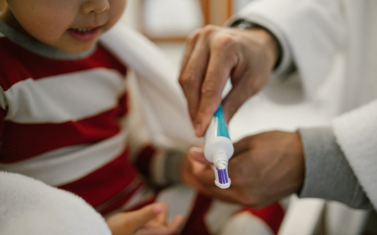 A close-up of a father helping his young daughter apply toothpaste on a toothbrush.