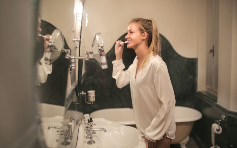 Side view of blonde female with ponytail in blouse standing in bathroom while brushing teeth and looking at mirror