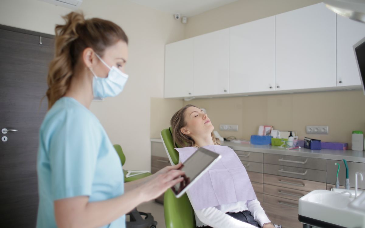 Female dentist in uniform and sterile mask touching tablet screen while standing near female patient sitting on medical
