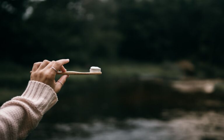 Hand holding a bamboo toothbrush with toothpaste against a natural outdoor backdrop.