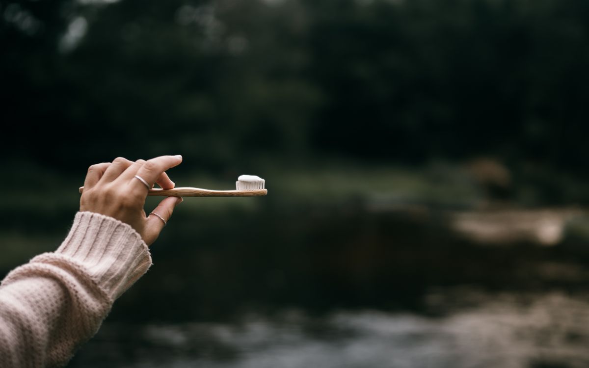 Hand holding a bamboo toothbrush with toothpaste against a natural outdoor backdrop.
