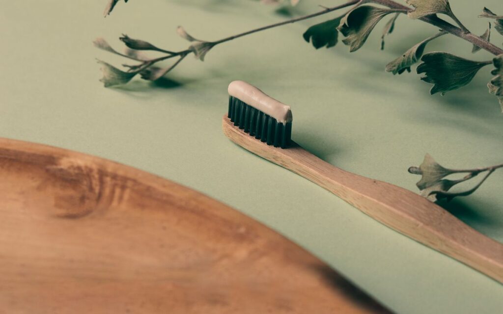 A bamboo toothbrush with toothpaste on a wooden tray beside green leaves, promoting natural oral care.
