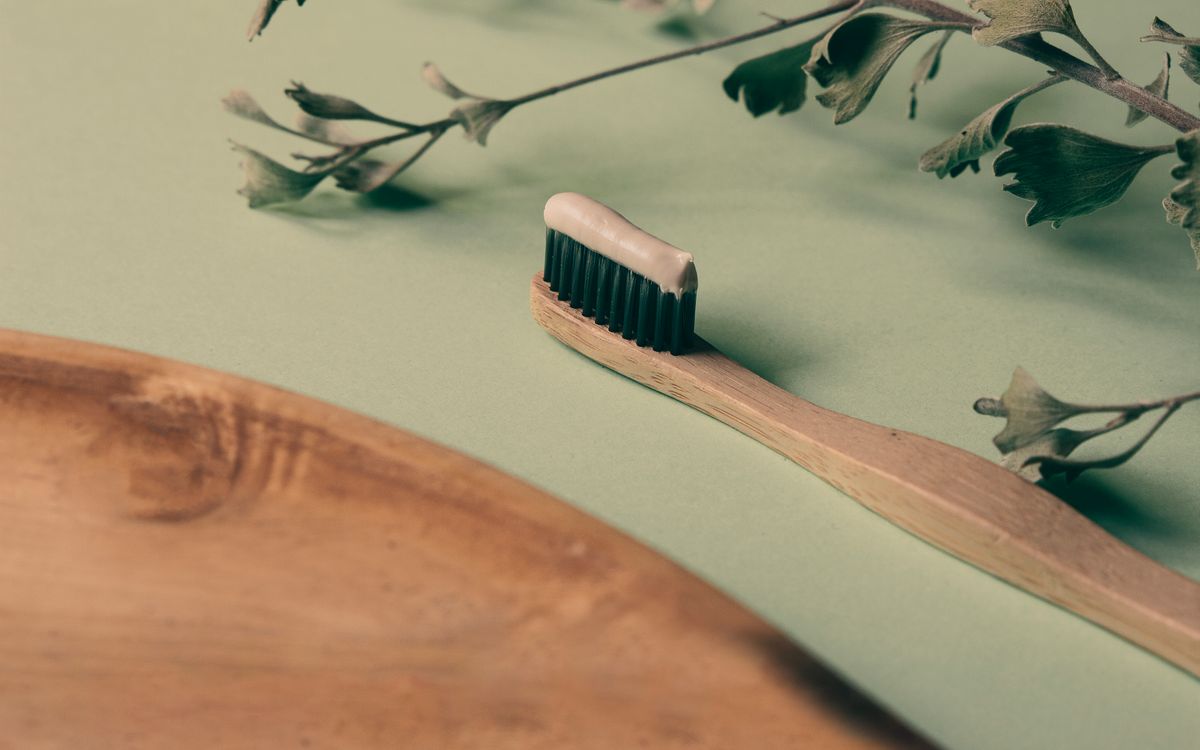 A bamboo toothbrush with toothpaste on a wooden tray beside green leaves, promoting natural oral care.