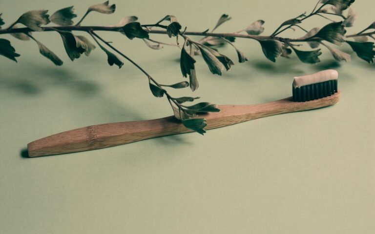 A bamboo toothbrush with toothpaste lies on a green background adorned with leaves, promoting natural oral care.